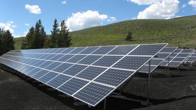 solar panels, green hill in the background with dark green christmas trees, below clear blue sky and beautiful clouds