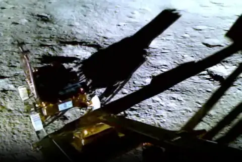 a gold colored small rover called pragyaan with white wheels travelling down a gold colored ramp of the vikram lander, on the rough silver colored surface of the moon